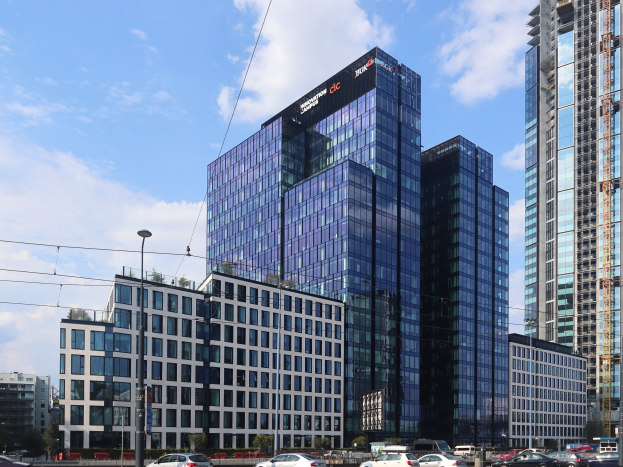 Außenansicht des Deutschen Bank-Hauptsitzes in Frankfurt, Deutschland, mit einem mehrstöckigen Gebäude mit zahlreichen Fenstern, umgeben von städtischer Infrastruktur wie Straßenmasten, Fahrzeugen und Grünflächen unter einem bewölkten Himmel.
